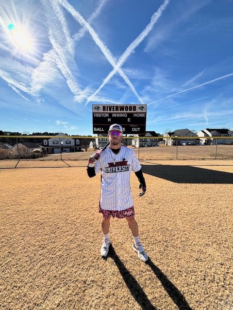 Person in a sports uniform standing on a dirt field with a sign and a blue sky with contrails behind.
