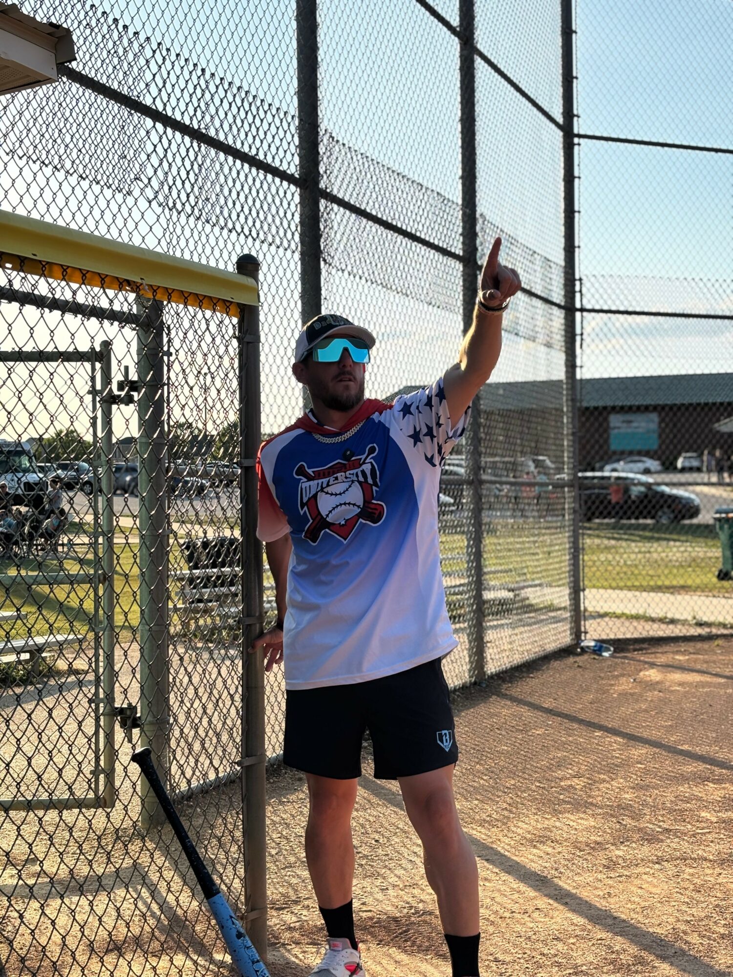 Person in baseball uniform and sunglasses pointing upward on a baseball field, with a chain-link fence and parking lot in background.