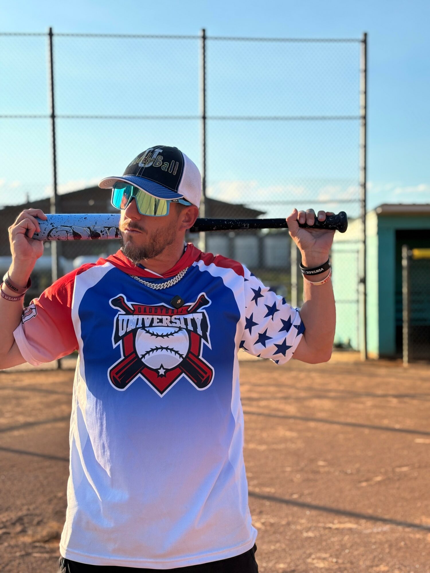 Baseball player holding bat over shoulder on dirt field, wearing jersey with stars and a logo, with a fence and building in background.