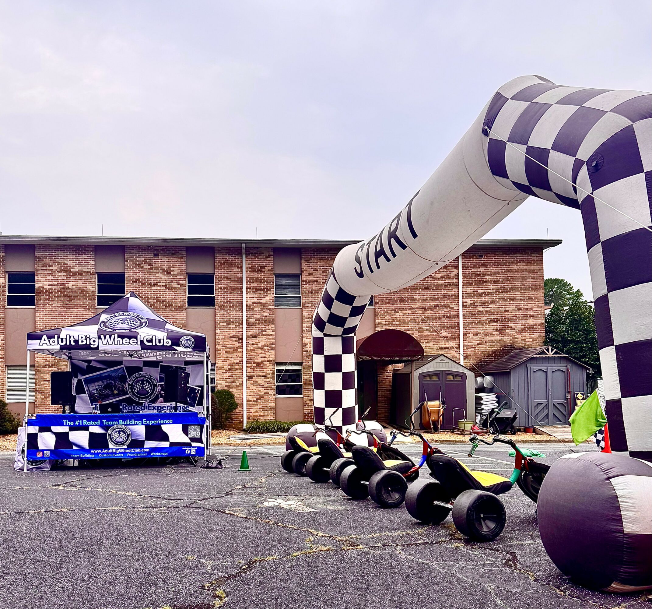 Outdoor event with a checkered arch, tires, and a vehicle in front of a brick building, with small structures and trees in background.