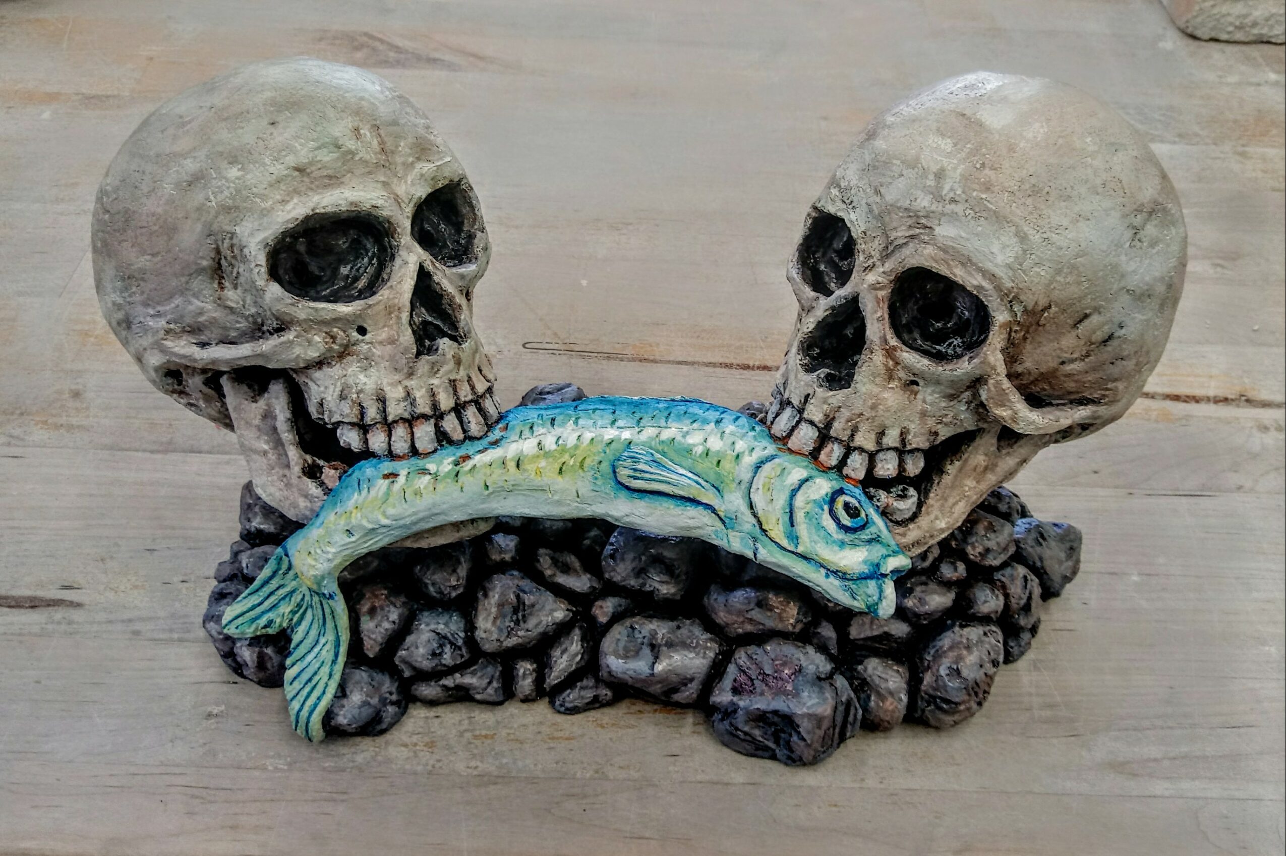 Decorative sculpture of two skulls with a fish between them, set on rocks, on a wooden surface.