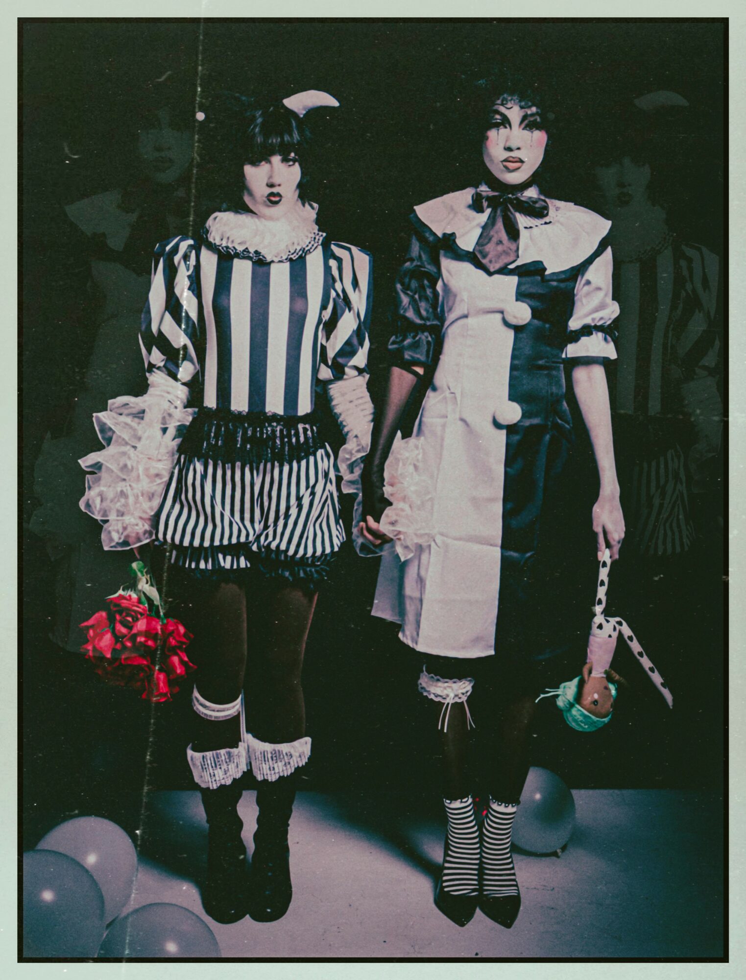 Two women in vintage-style costumes stand with balloons and flowers, one holding a small purse, in a dark setting.