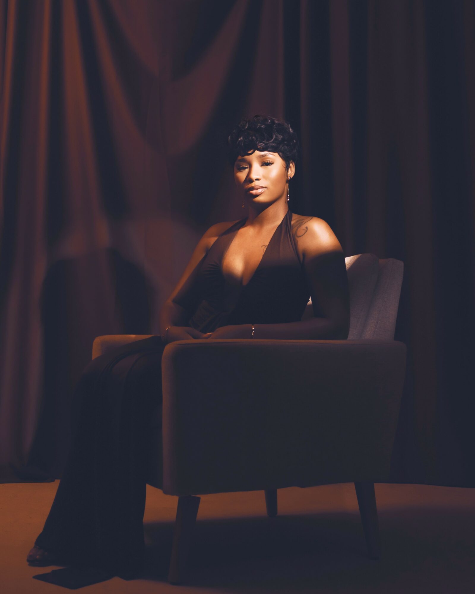 Woman with short curly hair sitting on a dark armchair against a dark curtain background.