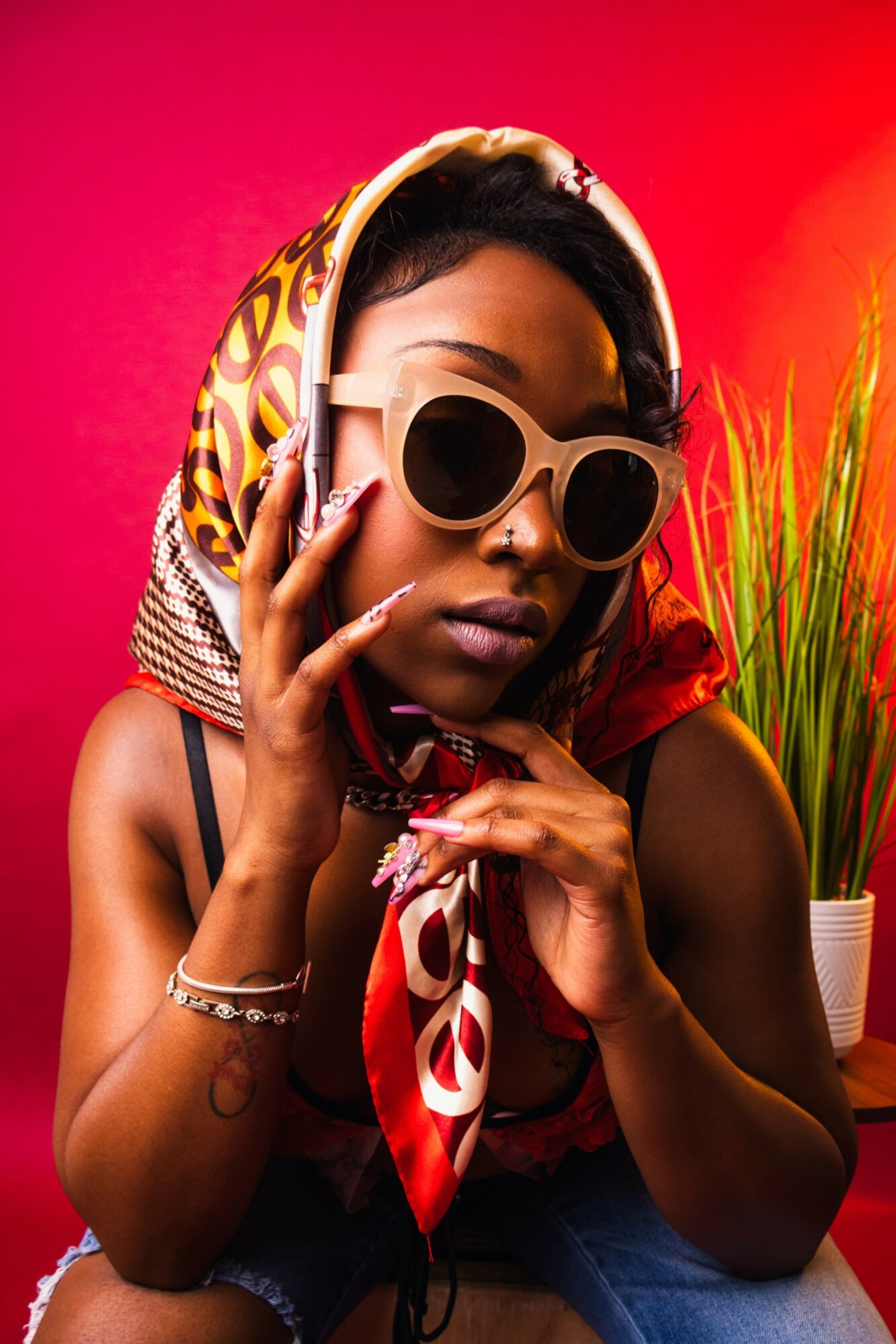 Woman with sunglasses and headscarf, posing indoors against a pink background, with a plant nearby.