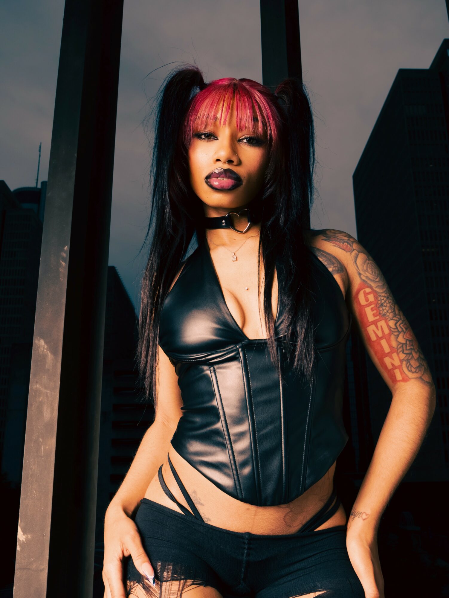 Woman with pink and black hair, wearing a black leather top, standing outdoors with city buildings in background.