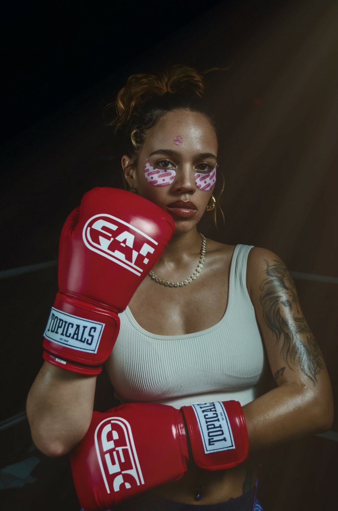 Woman with face paint, boxing gloves, and tattoos, looking serious, in a dark setting with light shining on her.