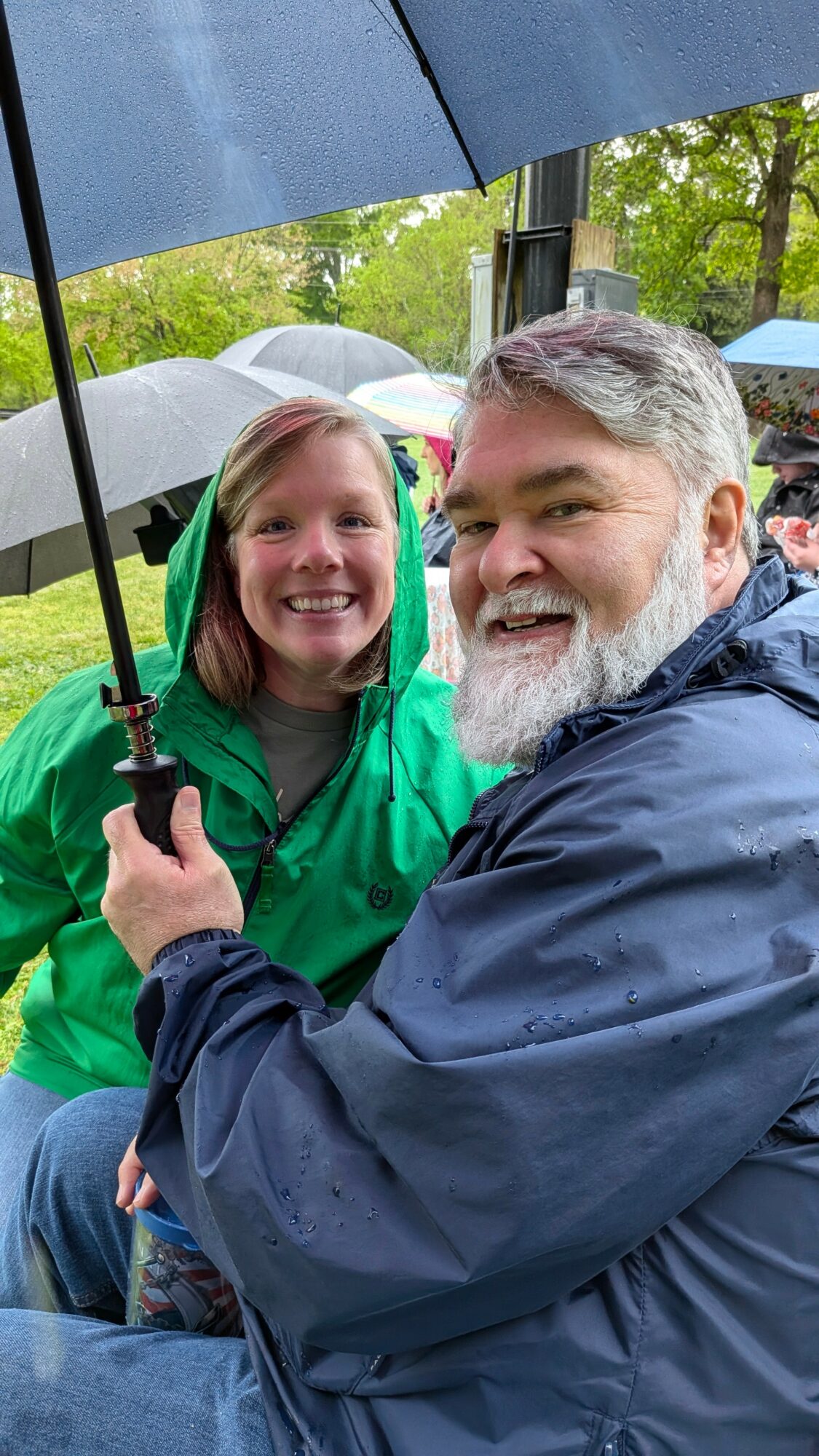 A man and a girl sit close together under umbrellas outdoors, smiling at the camera in a rainy setting.