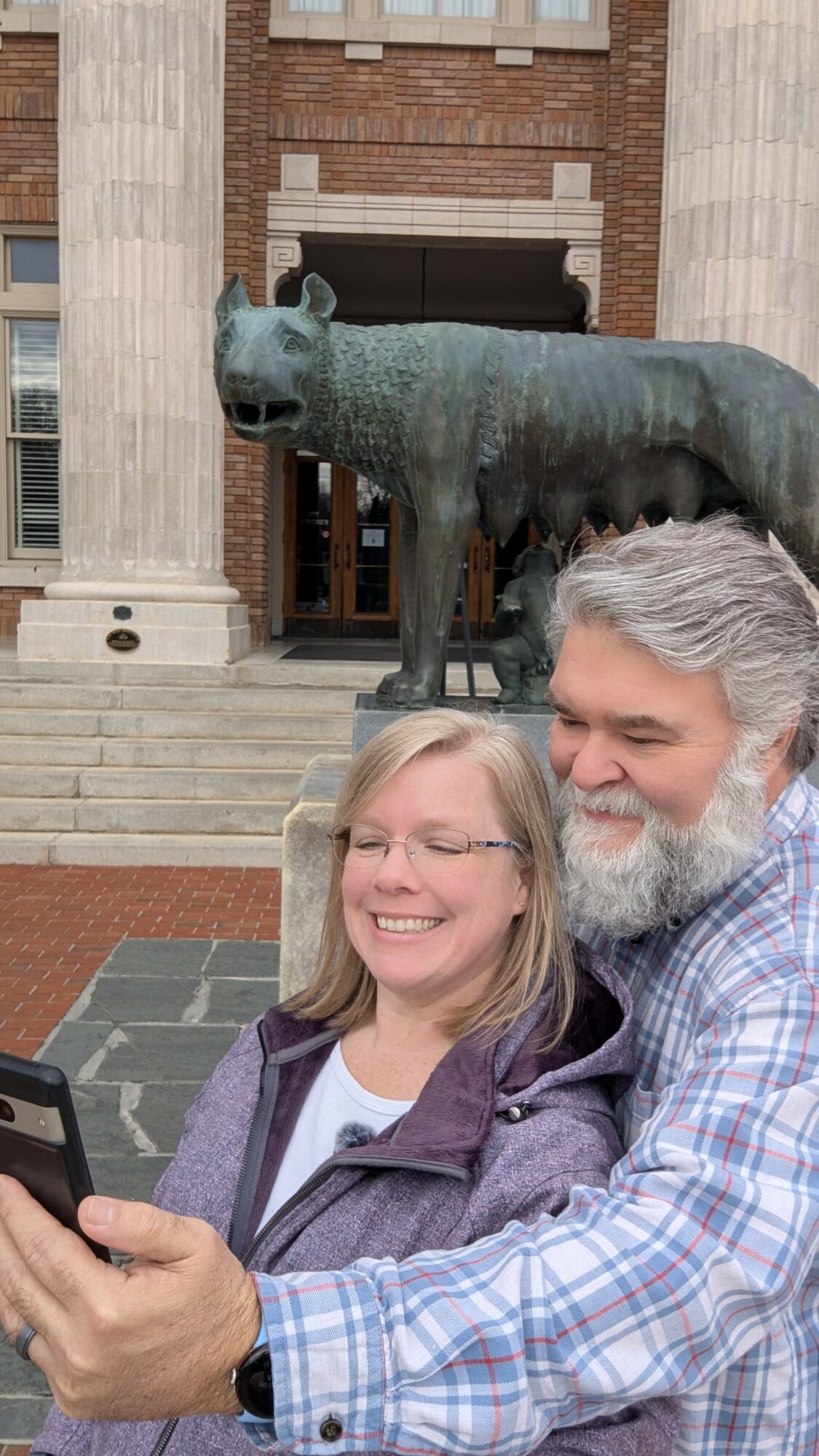 Two smiling people taking a selfie in front of a large bear statue outside a building with columns.