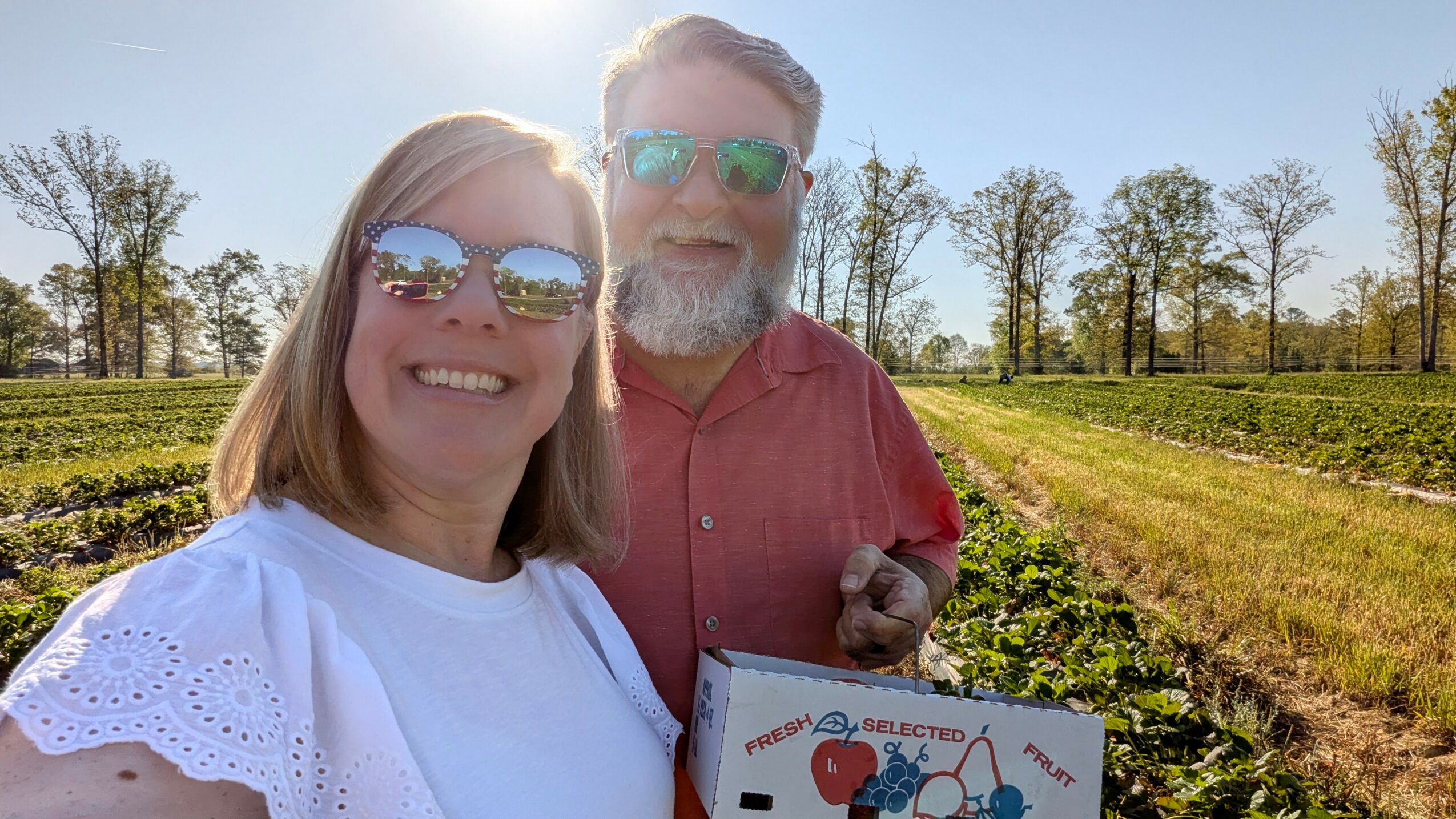 Two people smiling outdoors, holding a box, with trees and fields in the background.