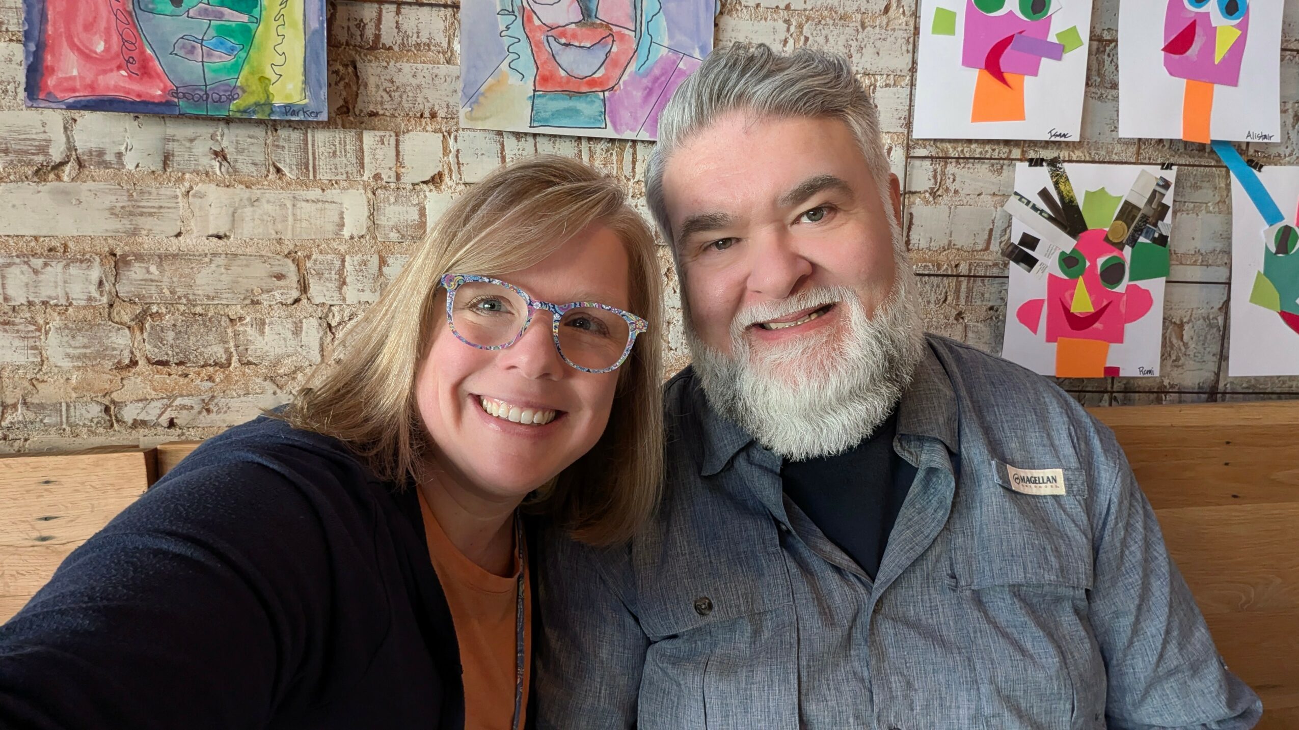Two people smiling, one with glasses and a beard, in front of colorful artwork on a brick wall.