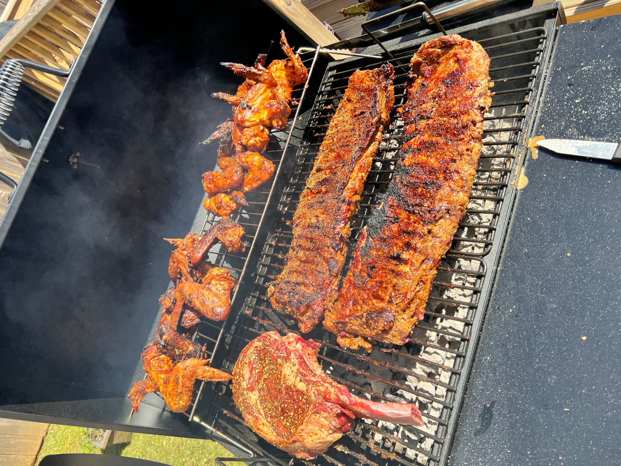 Barbecue grill with skewered chicken, two large seasoned steaks, and a piece of meat with a bone, cooking outdoors.