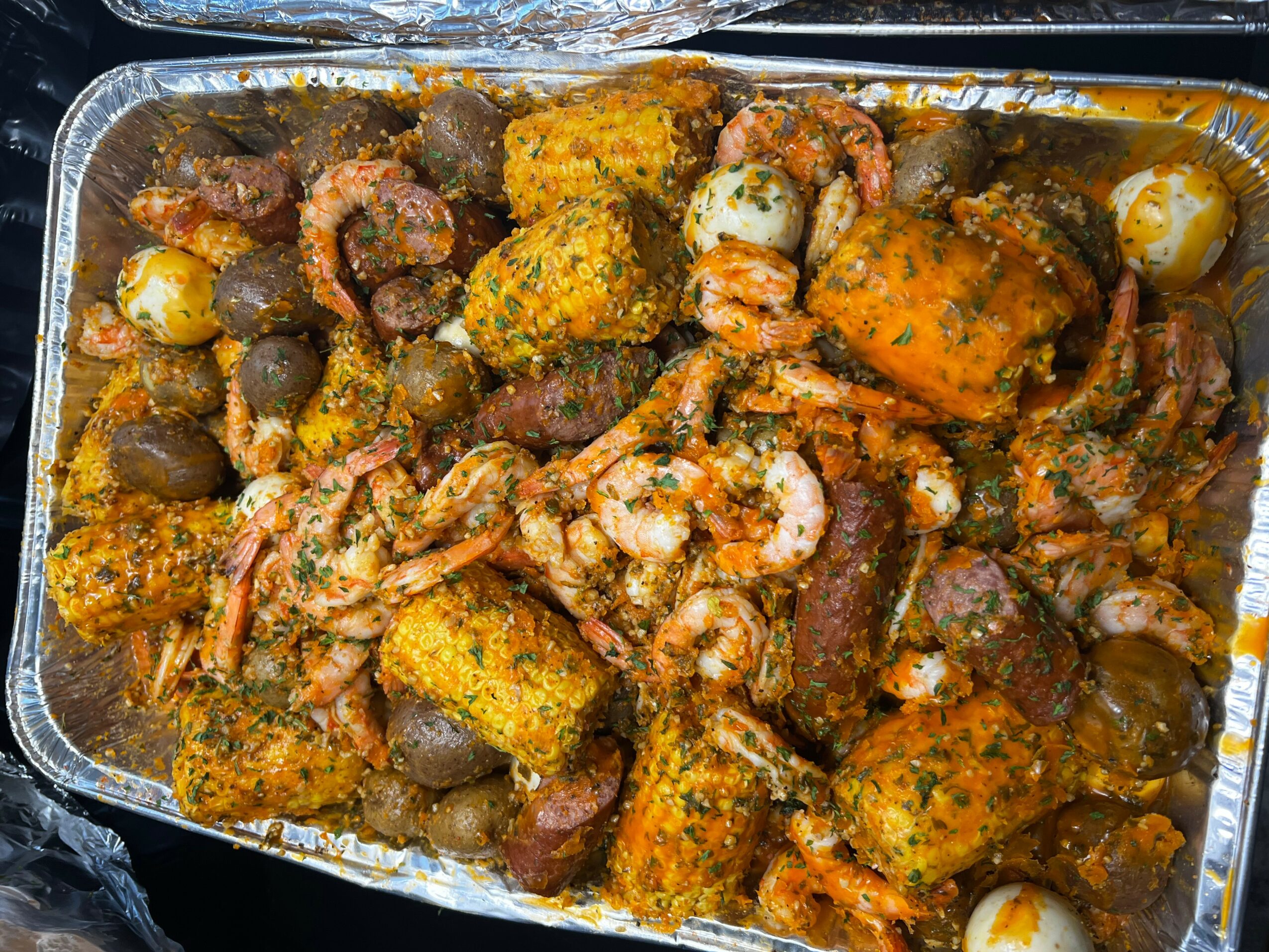 Assorted cooked seafood and vegetables in a metal tray, including shrimp, corn, and potatoes, seasoned with spices.