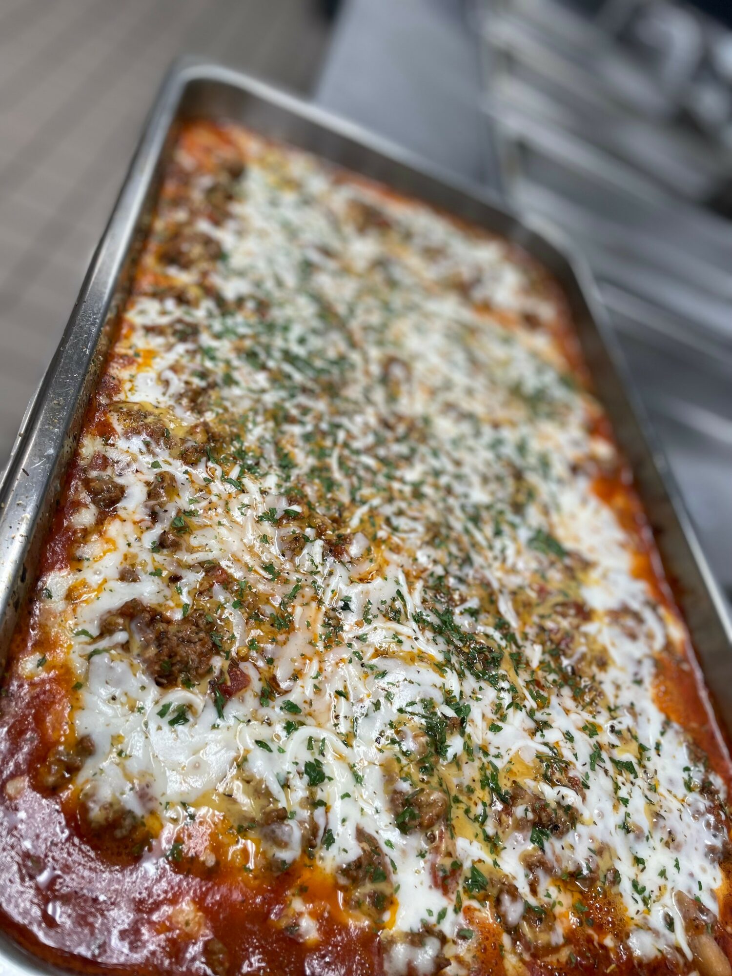 Rectangular tray of baked lasagna with cheese and herbs, viewed from the side, on a kitchen surface.
