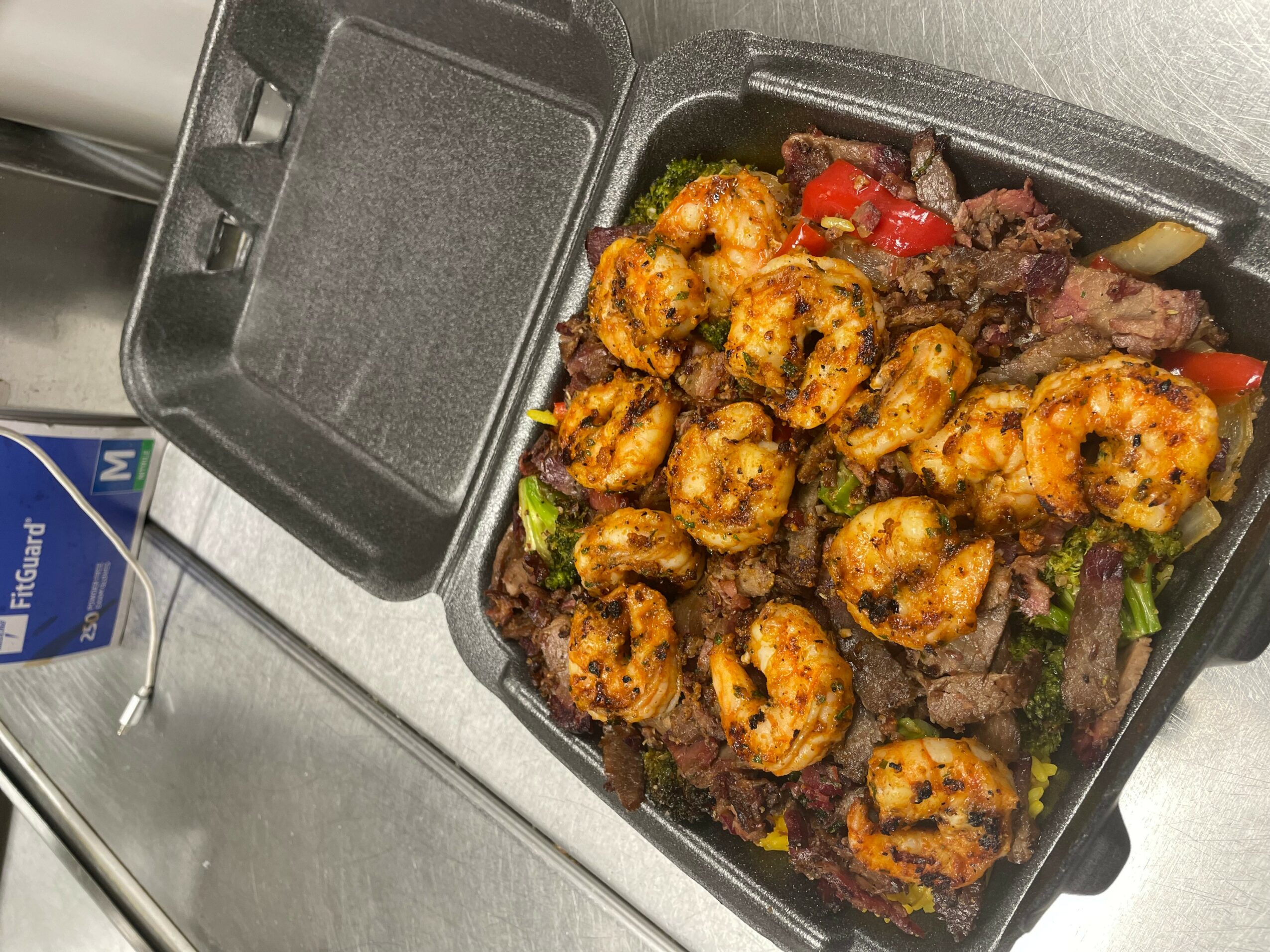 Shrimp and beef stir-fry in a black takeout container with vegetables, on a metal surface.