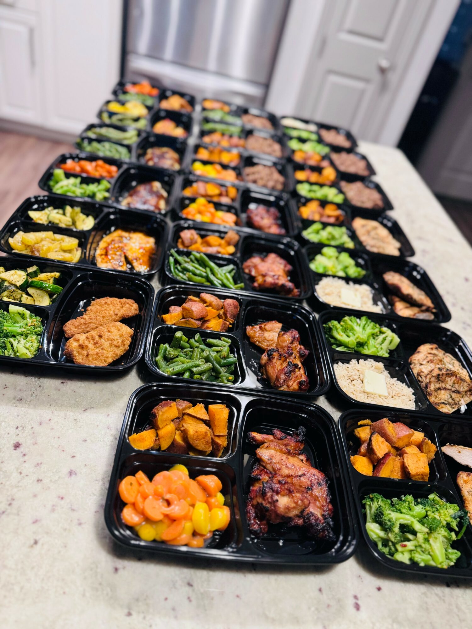 Multiple black meal prep containers with various cooked foods on a kitchen countertop, arranged in rows.