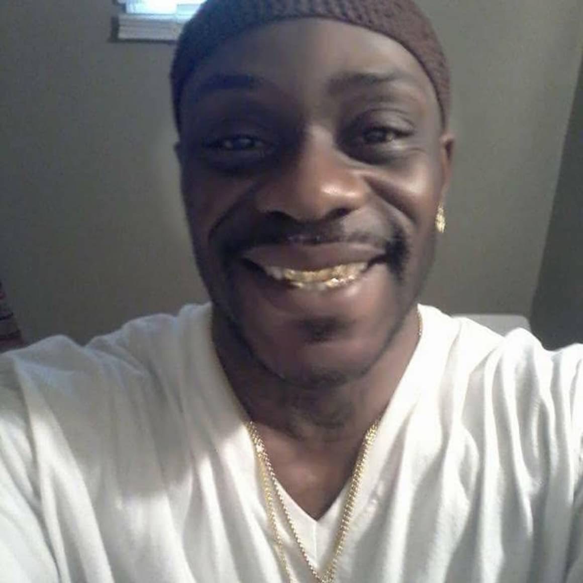 Smiling person with gold teeth, wearing a white shirt, gold chain, and a brown headband, indoors.
