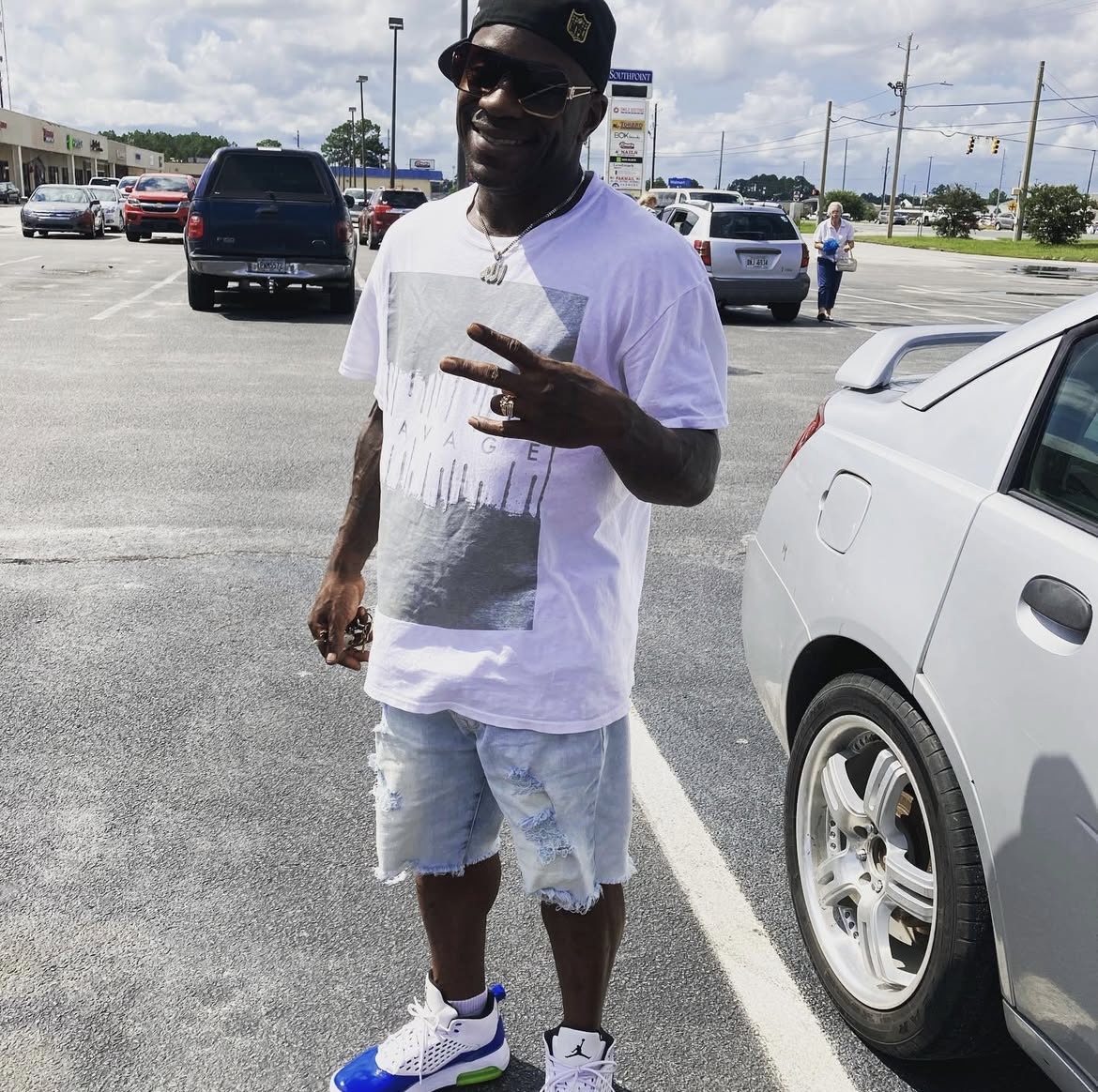 Person standing in parking lot, smiling, wearing a white t-shirt, denim shorts, sneakers, and a cap, making a peace sign.
