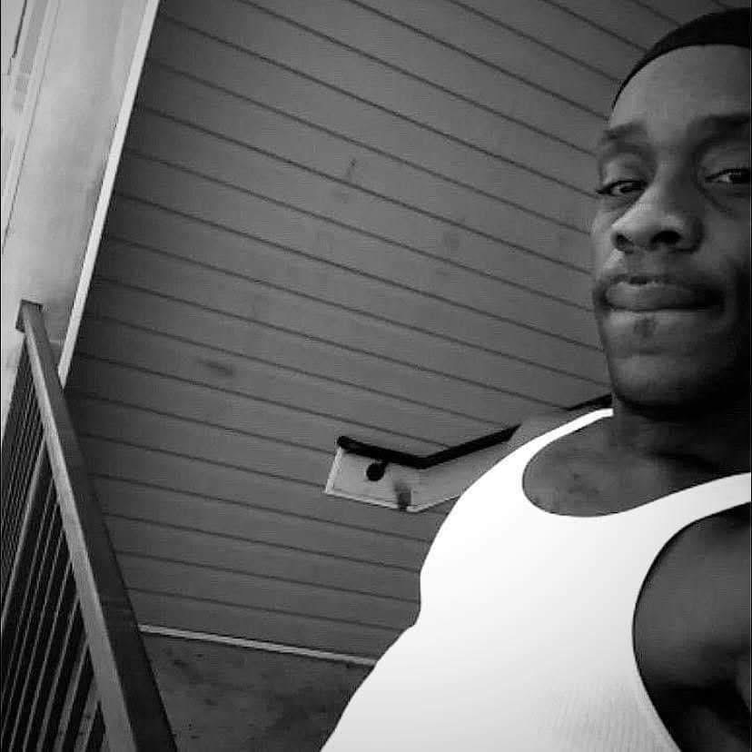 A man taking a selfie on a porch with horizontal siding, wearing a white tank top, smiling slightly.