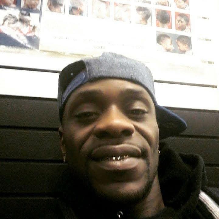 Young man wearing a gray cap backwards, smiling, with photos on the wall behind him.