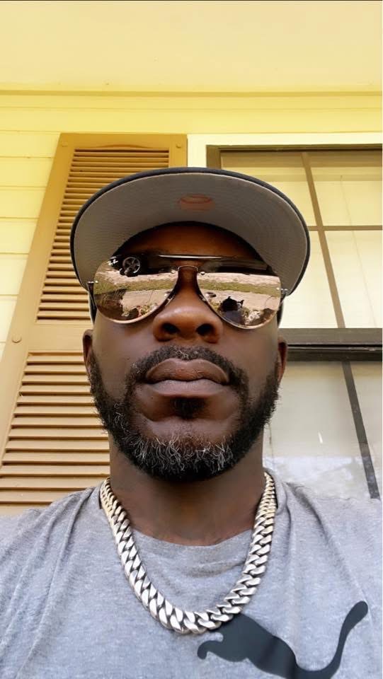 Man wearing sunglasses, cap, and chain, standing outside near a building with window and vent, looking at camera.
