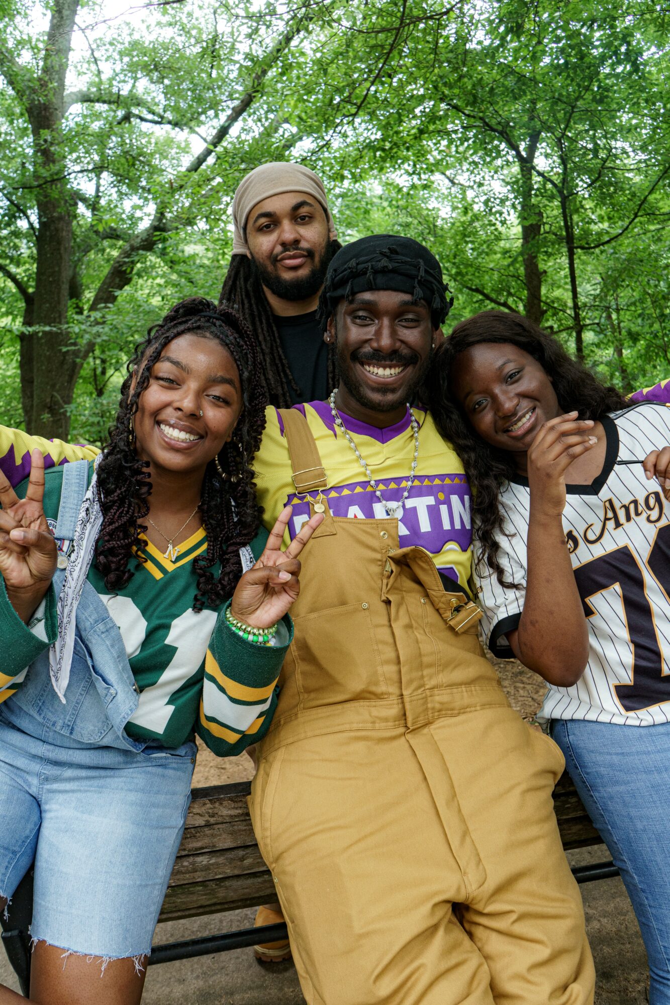 Four people smiling outdoors among green trees, two women and two men, one woman making a peace sign, one woman resting her head.