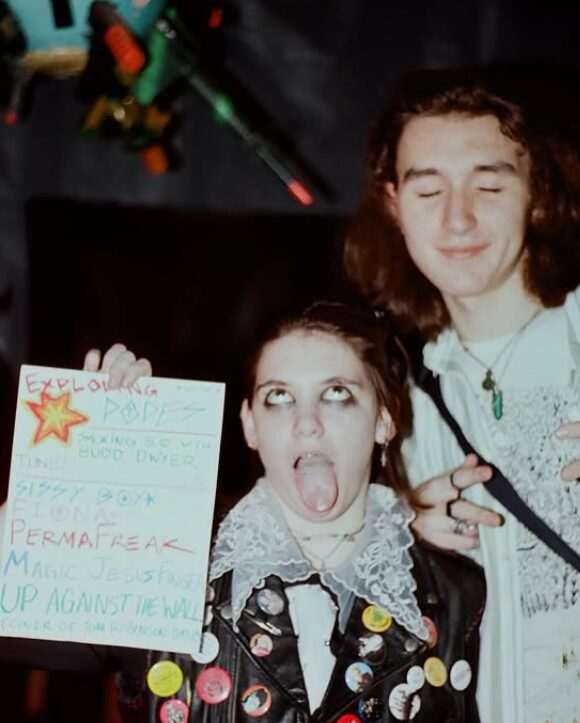 Two young women, one with eyes closed and smiling, the other with tongue out holding a sign, in a dark setting.