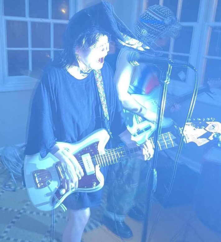 Two people playing electric guitars, one singing into a microphone, in a room with large windows, blue lighting.