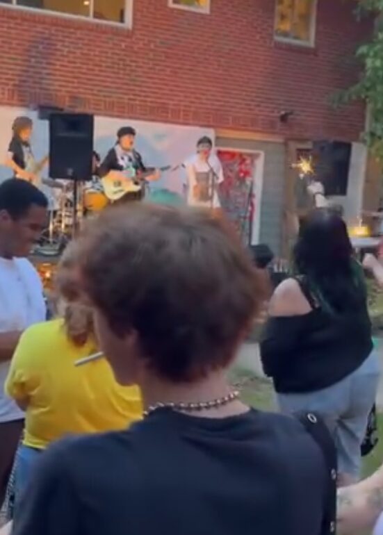 People watching a live outdoor music performance with a brick building in the background.