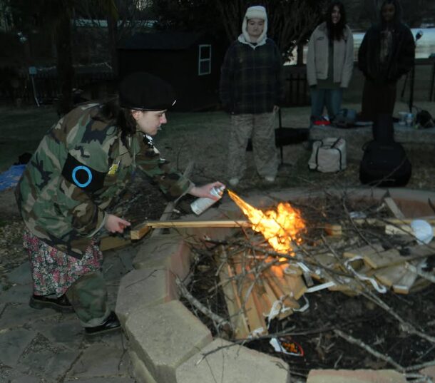 Person in camouflage jacket and black hat using a torch to ignite a fire in a fire pit, with four people watching nearby.