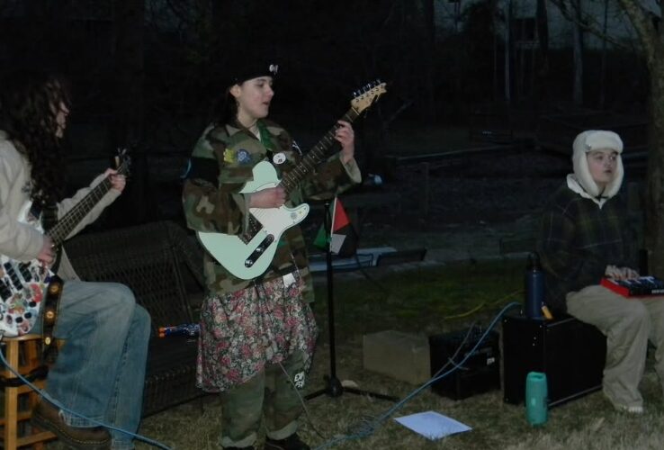 Three people playing guitars and keyboard outdoors at night, with trees and dark sky in background.