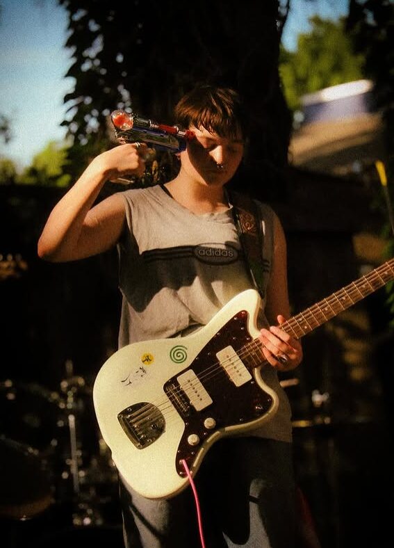Child holding a toy gun to their head and playing an electric guitar outdoors, with trees and sky in background.
