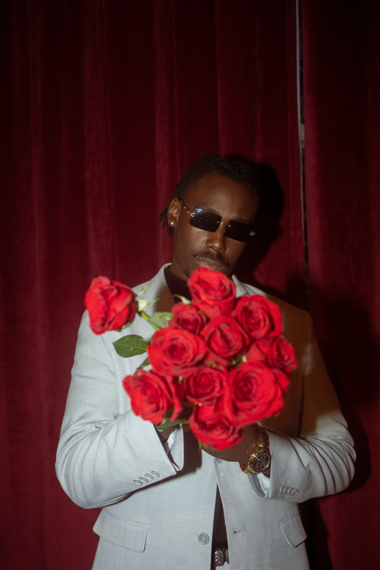 Person with sunglasses holding a large bouquet of red roses, standing in front of a red curtain.