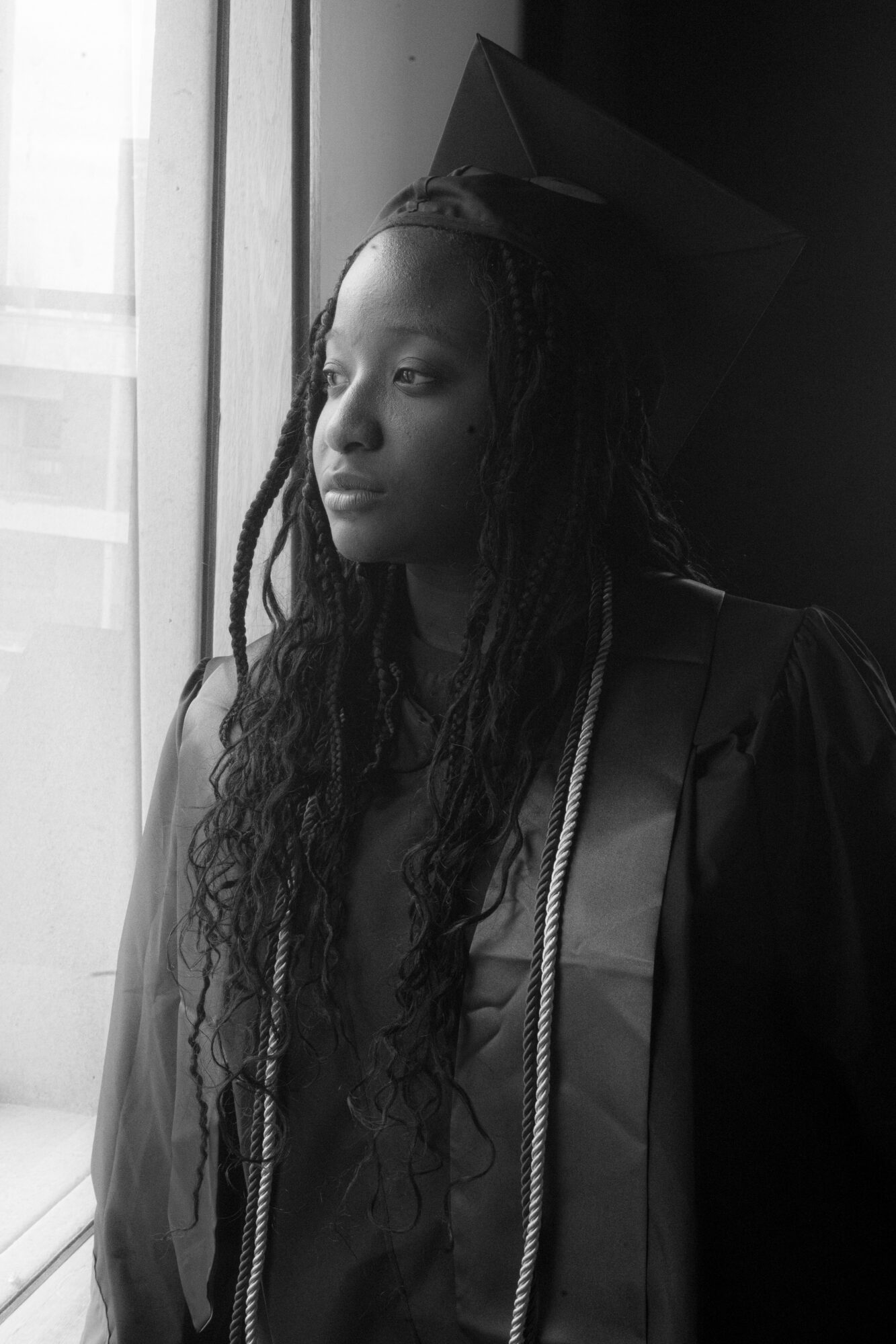 Young woman in graduation cap and gown looking out window, with long curly hair, in black and white.