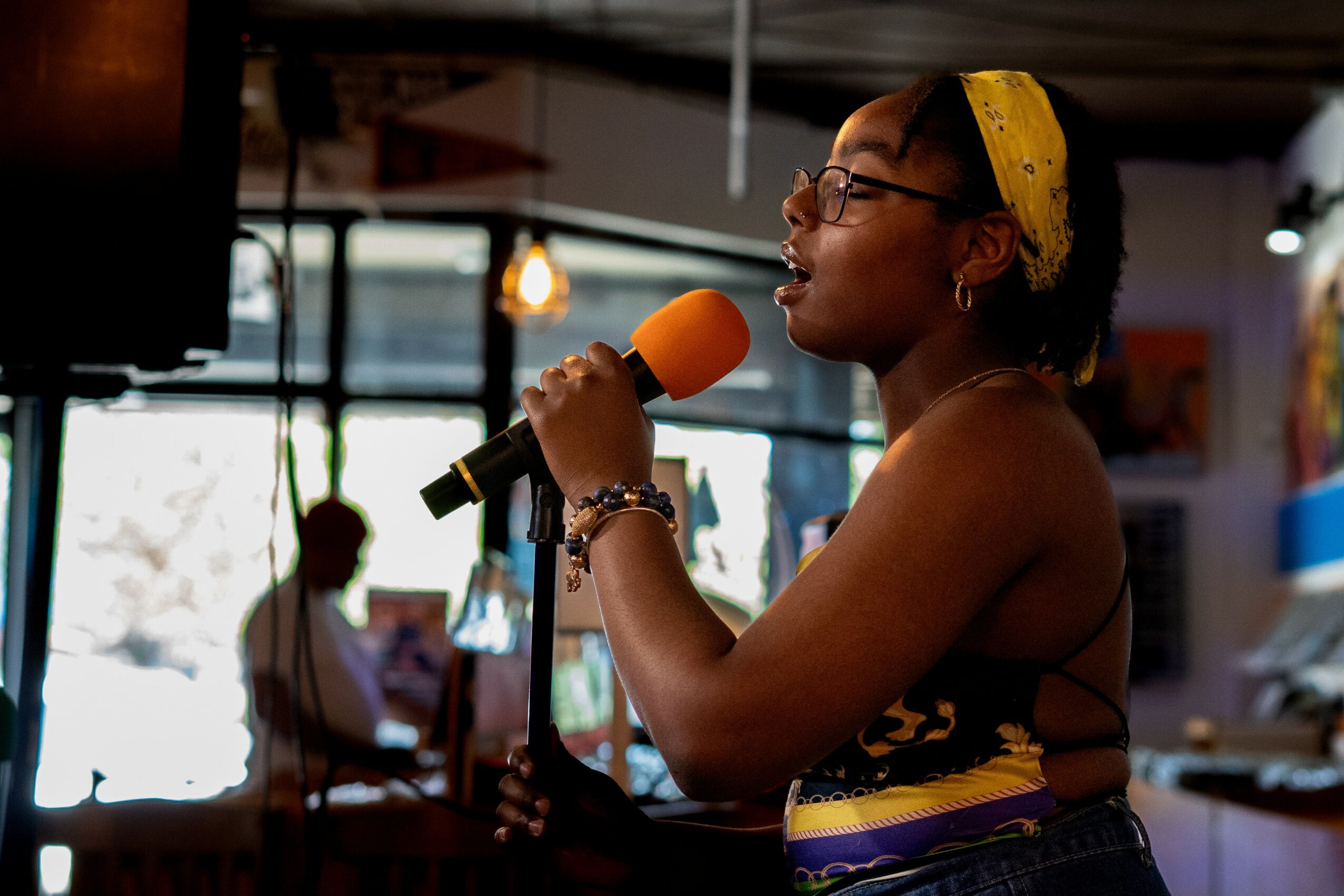 Woman singing into a microphone with a yellow headband in a cafe, with people and windows in the background.