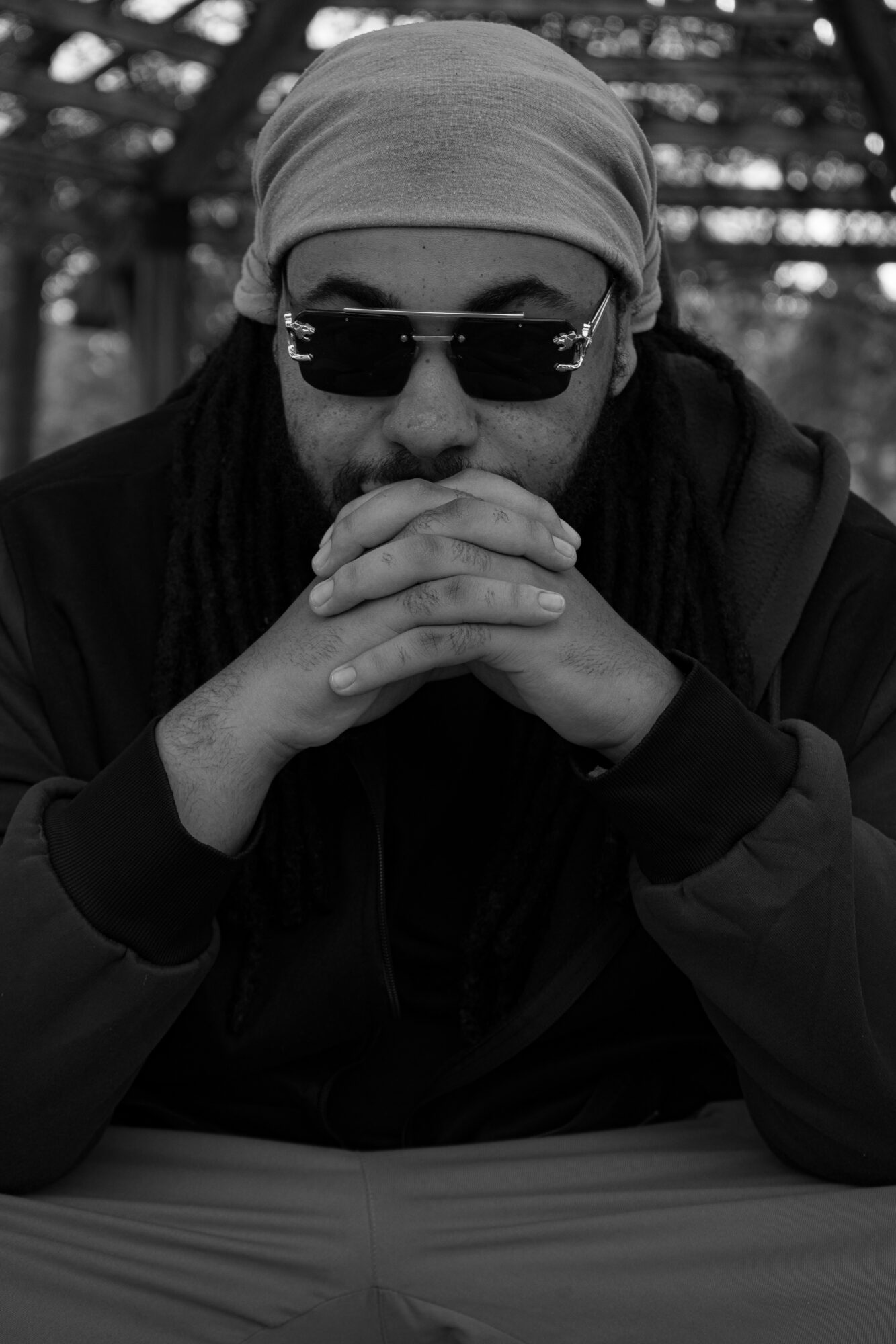 Person wearing sunglasses and a headscarf, resting hands together, outdoors with trees in background.
