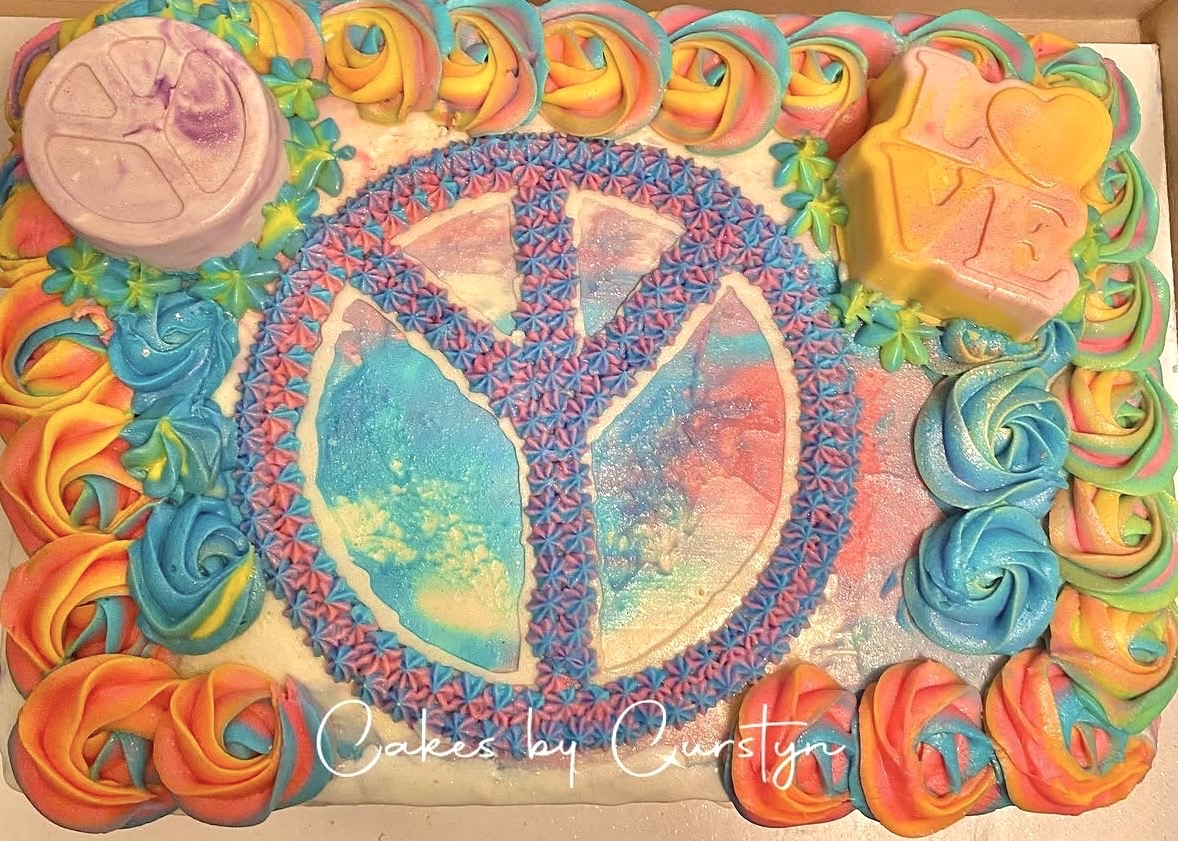 Colorful cake with peace symbol, decorated with rainbow-colored frosting and various shaped decorations, including a pink circle and yellow star.
