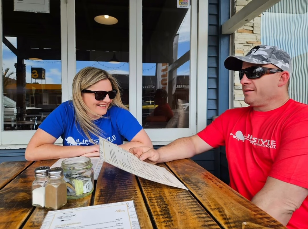Two people sit at a wooden table outside a cafe, looking at a menu, with jars on the table.
