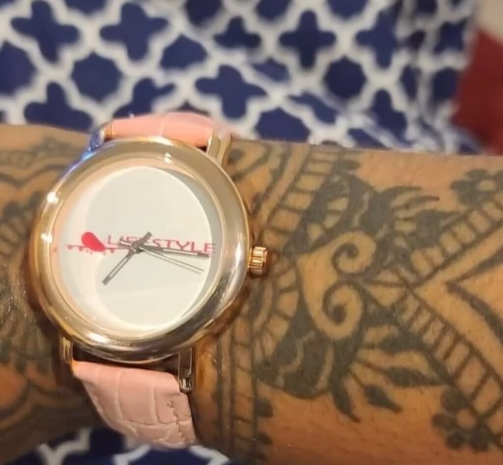 Person's arm with a pink watch, tattooed skin, and a patterned shirt in the background.