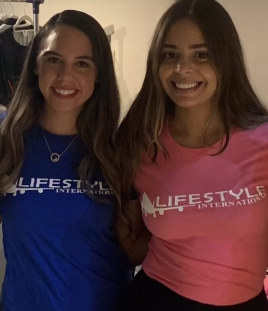 Two women smiling, standing close, wearing T-shirts with 'LIFESTYLE' text, one in blue, one in pink.