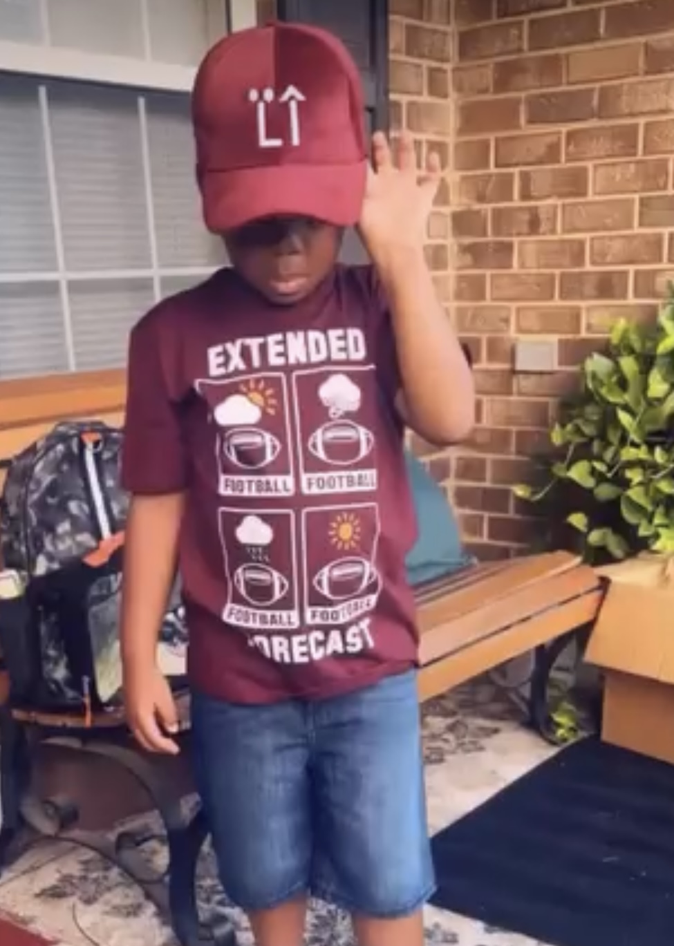 Child wearing a red cap and maroon T-shirt standing outdoors near a brick wall and bench.