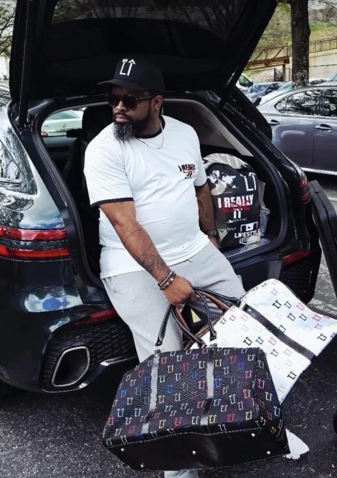 Man with sunglasses and beard loading bags into the trunk of a black car, wearing a white t-shirt and cap.