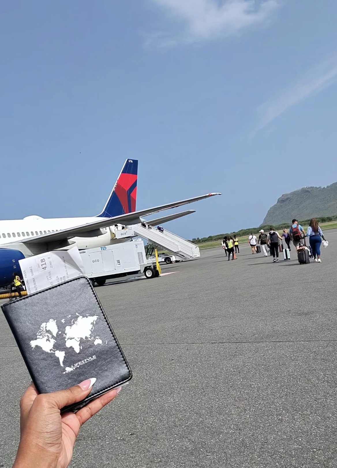 Person holding a world map passport in front of an airplane on the tarmac with people walking nearby, clear sky above.