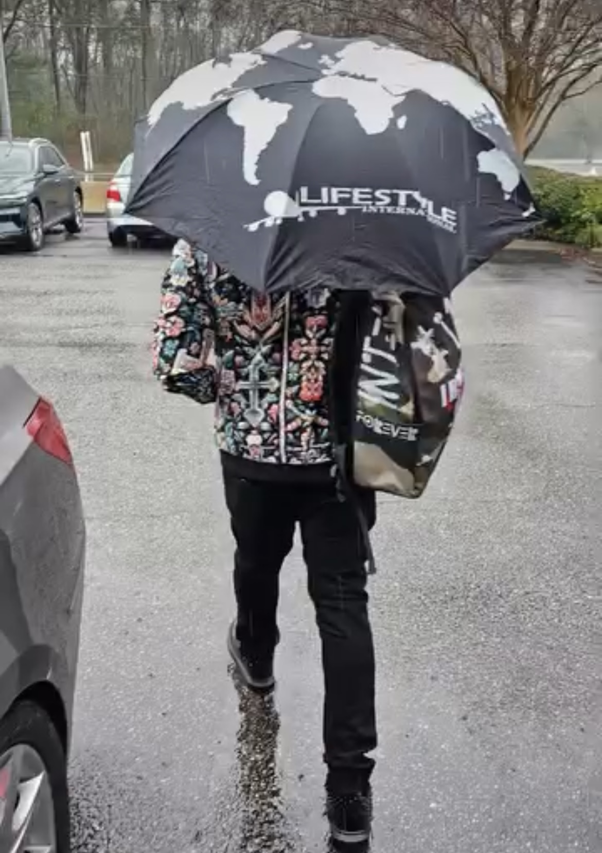 Person walking on wet pavement holding a large umbrella with a world map design, wearing a floral jacket and carrying a bag.