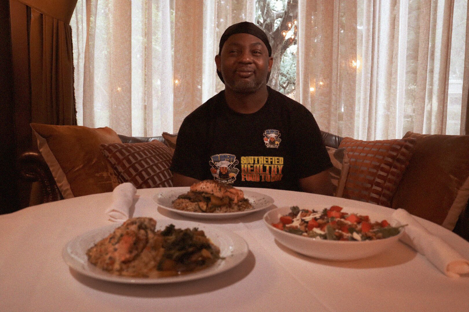 Person sitting at a table with three plates of food, in a room with curtains and cushions behind.