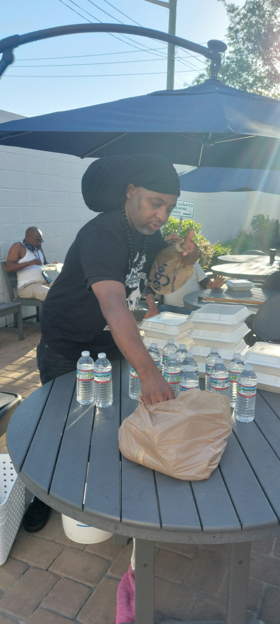 Person arranging bottled water on a round table outdoors under a large umbrella, with other people in the background.