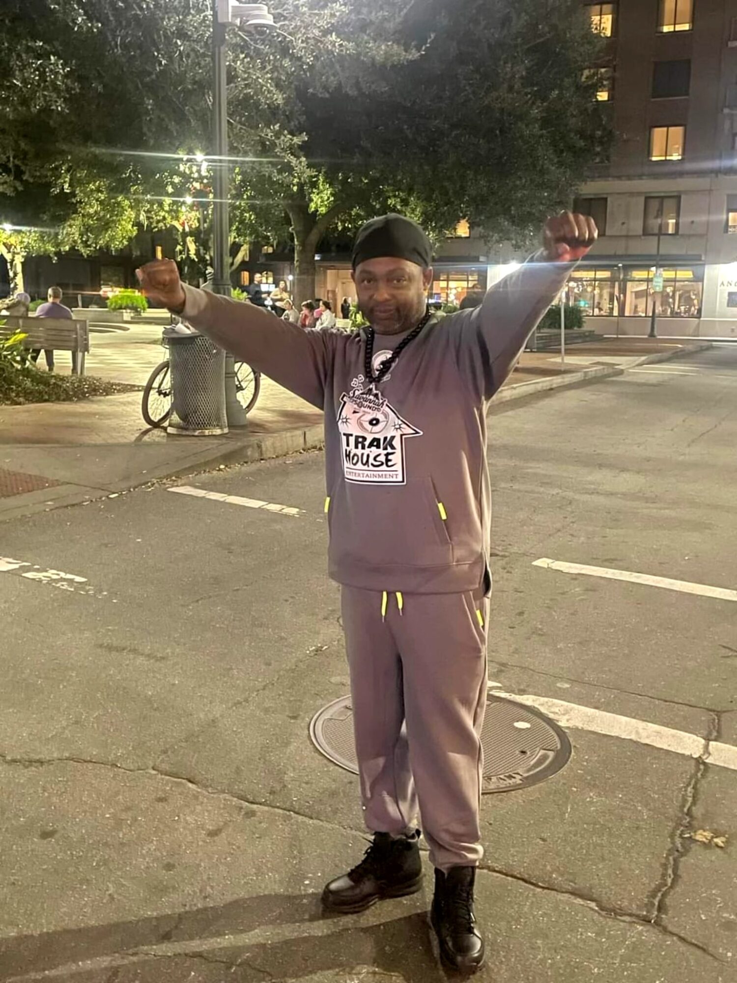 Person standing outdoors with arms raised, smiling, in a city setting at night, trees and buildings in background.