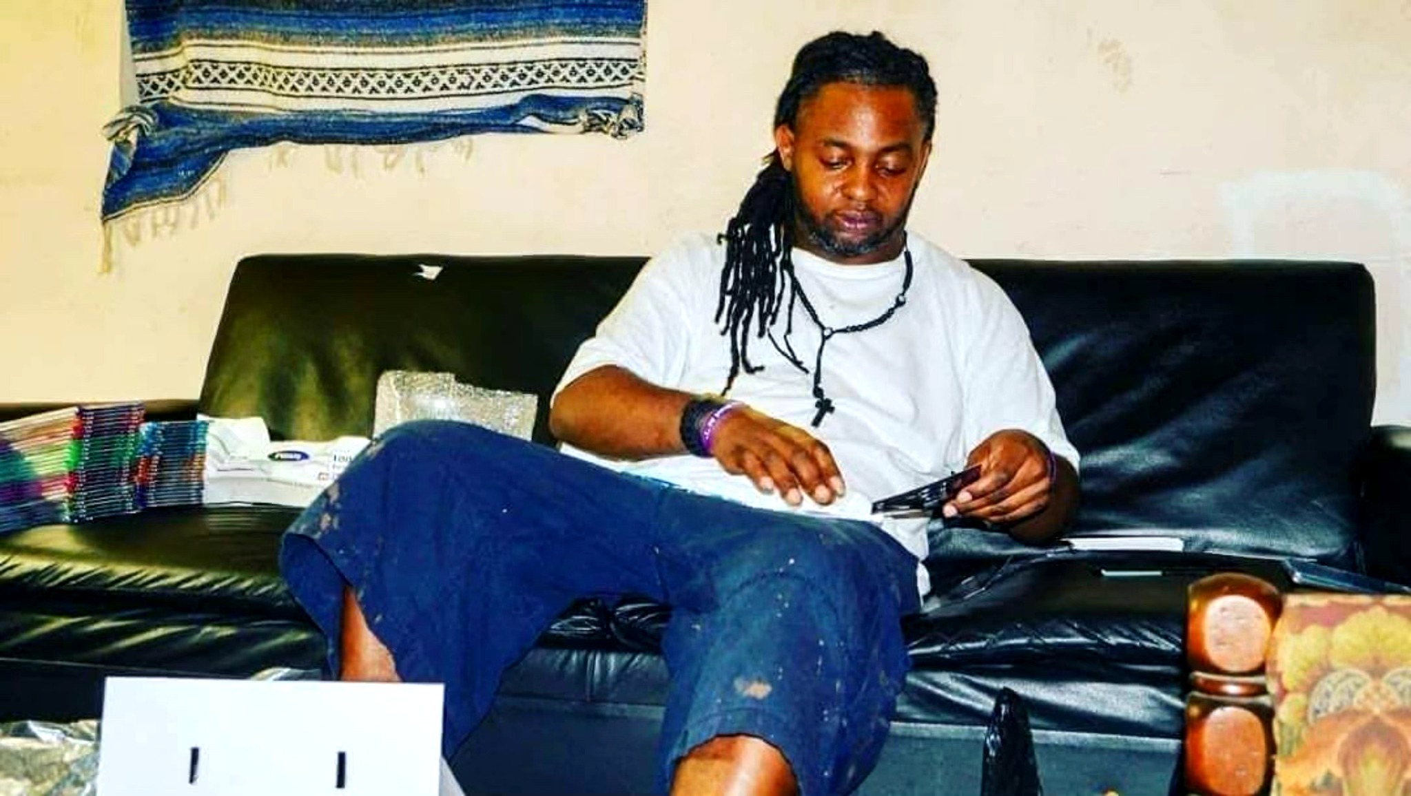 Person with long dreadlocks sitting on a black sofa, using a smartphone, with a window and various objects nearby.