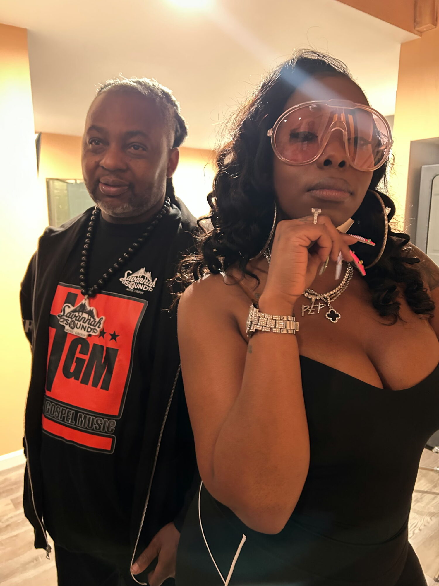 A woman with large pink sunglasses and a man with dreadlocks pose indoors, both looking at the camera.