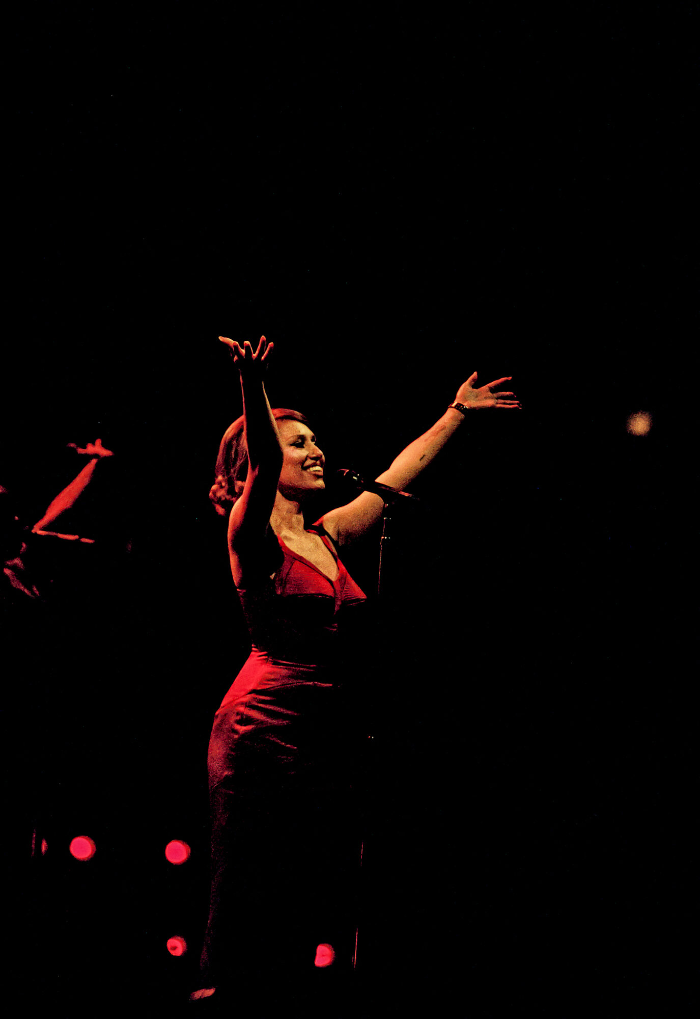 A woman in a red dress is performing on stage with her arms raised, against a dark background with stage lights.