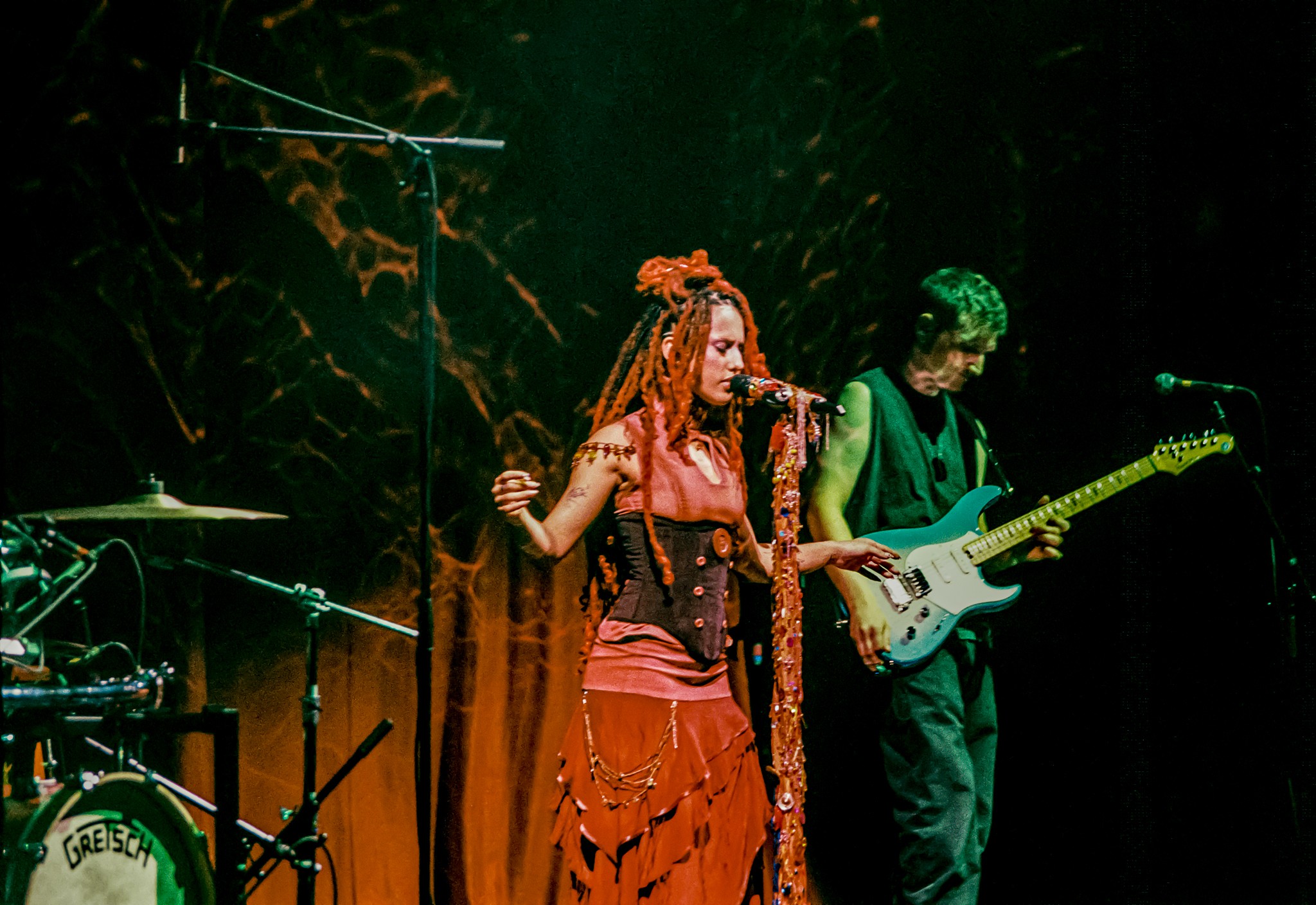 Two musicians perform on stage, one singing with long hair and colorful clothing, the other playing guitar, with stage lighting.